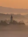 Church tower and rooftops emerging from mist at sunrise in a hilly countryside.