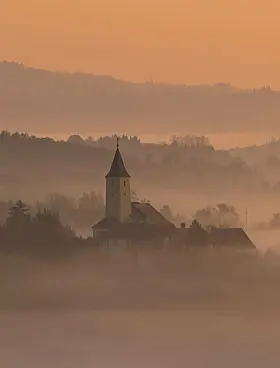 Church tower and rooftops emerging from mist at sunrise in a hilly countryside.