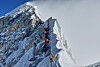 A line of climbers in orange gear ascending a snowy ridge on Mount Everest, fixed ropes visible.