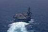 Aerial view of USS George H.W. Bush aircraft carrier moving through open sea, leaving white wake.