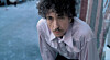 Bob Dylan med vådt hår og lys skjorte står ved en mur med blå baggrund.