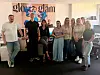 En gruppe står samlet foran en plakat med teksten «glo glam» i et lyst rom.