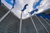 European Union flags on flagpoles in front of the curved glass facade of the Berlaymont building in Brussels, photographed from below against a cloudy sky.