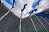 European Union flags on flagpoles in front of the curved glass facade of the Berlaymont building in Brussels, photographed from below against a cloudy sky.