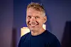 Smiling man in a dark shirt posing against a blue and purple studio background
