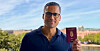 A man wearing glasses holds a burgundy Danish passport outdoors, with a waterfront cityscape behind him.