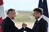Two suited men shake hands between Canadian and French flags outdoors.